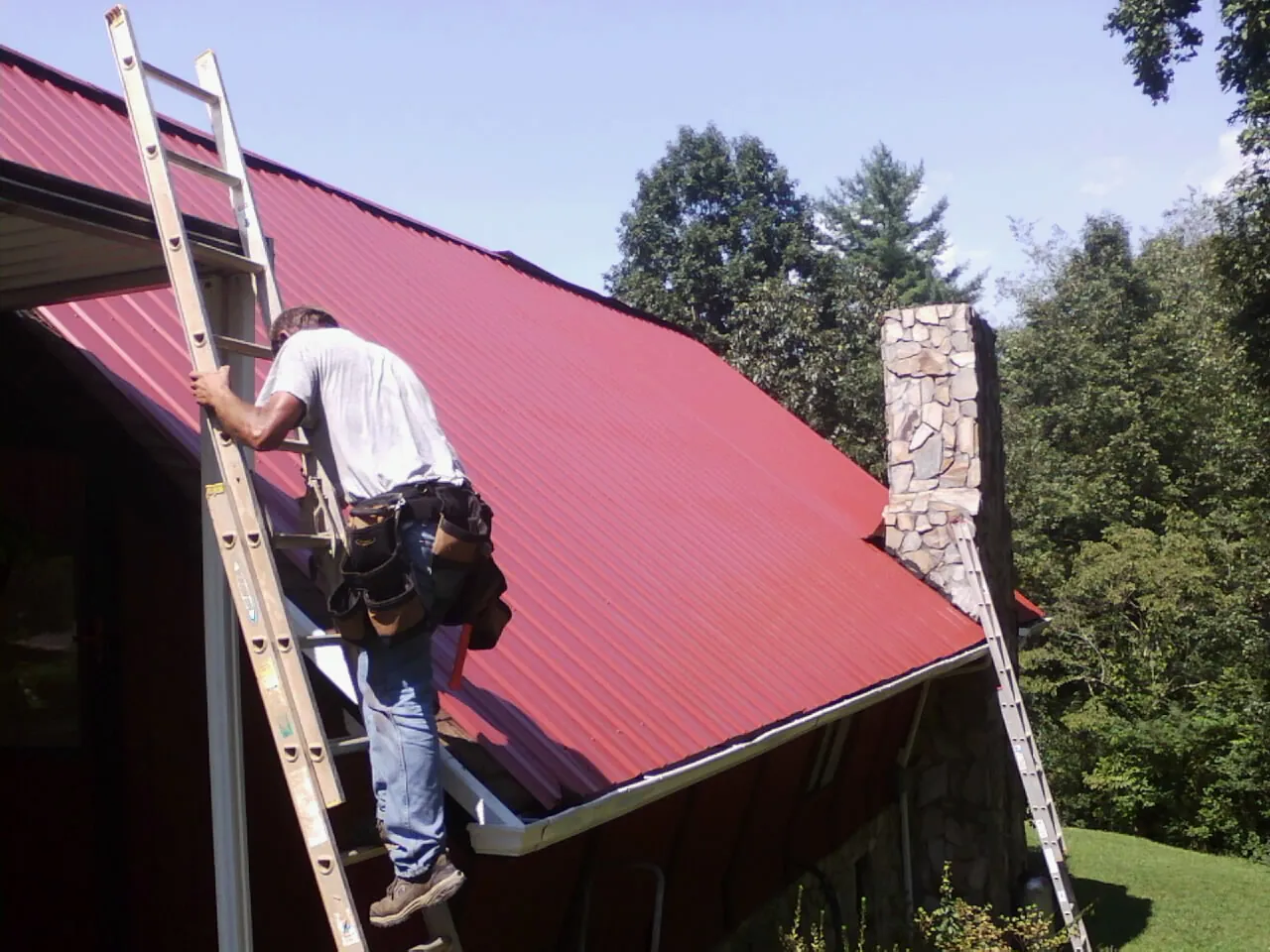 Roofer on a ladder beside a red metal roof and stone chimney — Soffit & Fascia Repair by Roofing Brothers in New Zion